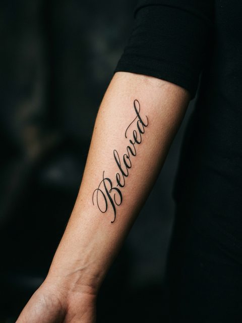 The word Beloved in flowing black script tattooed along a forearm